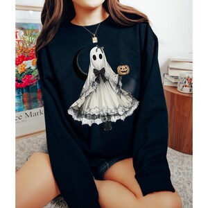 以下が含まれることがあります： 白い幽霊がジャック・オ・ランタンを持っている黒いスウェットシャツ。幽霊はレースのトリムと黒いリボンが付いた白いドレスを着ています。幽霊は三日月の前に立っています。