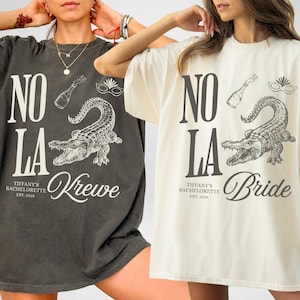 Two oversized t-shirts, one pepper and one ivory, featuring a graphic design. The design includes the text "NO LA", an alligator, a champagne bottle, and the text "Tiffany's Bachelorette Est. 2025" and "Krewe" or "Bride".