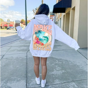 May include: White hoodie with the words "MAKE WAVES" in a retro font. The back graphic features a tropical scene with palm trees, waves, and flowers. The hoodie is worn by a person on a sidewalk.