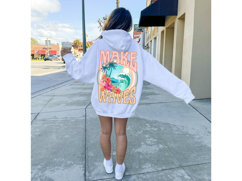 May include: White hoodie with the words "MAKE WAVES" in a retro font. The back graphic features a tropical scene with palm trees, waves, and flowers. The hoodie is worn by a person on a sidewalk.