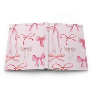 May include: Open journal with a light pink cover and pages. The cover features a repeating pattern of watercolor bows in shades of pink and beige. The journal is open, displaying the interior pages.