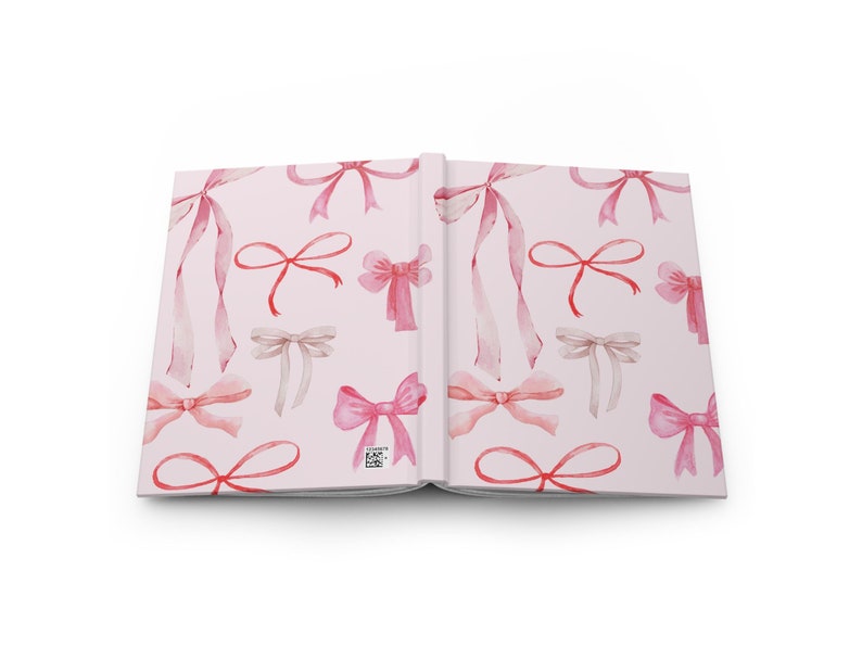 May include: Open journal with a light pink cover and pages. The cover features a repeating pattern of watercolor bows in shades of pink and beige. The journal is open, displaying the interior pages.