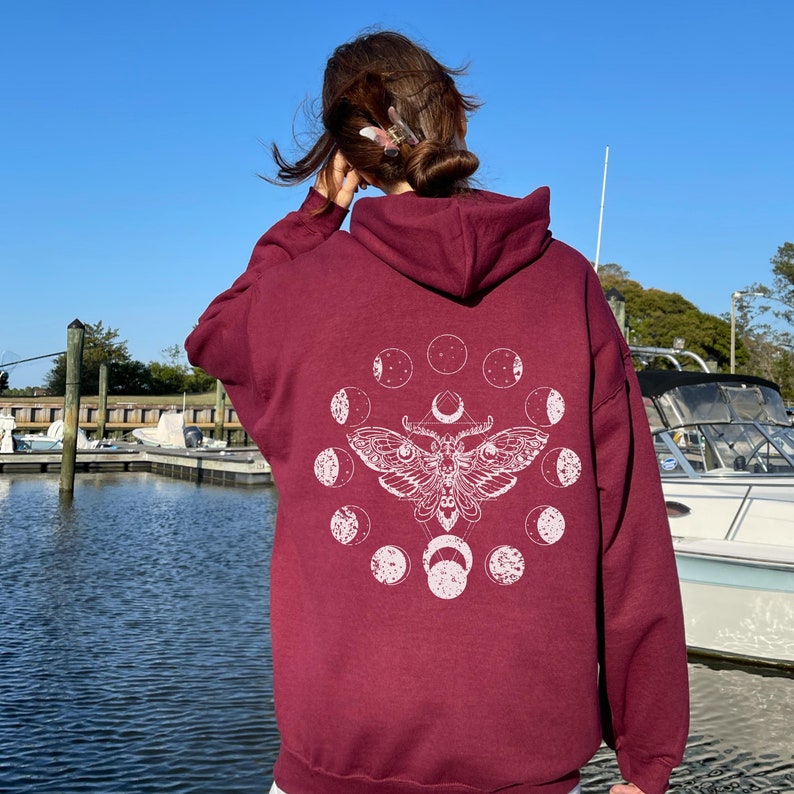 May include: A maroon hoodie with a white moth and moon phase design on the back. The graphic features a detailed moth illustration surrounded by a circle of moon phases. The hoodie has a hood and a relaxed fit, perfect for casual wear.