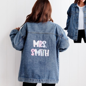 Puede incluir: Chaqueta vaquera azul claro con el texto "MRS. SMITH" en un diseño floral en la espalda. La chaqueta tiene un diseño clásico con botones y mangas largas. La imagen muestra a una persona que lleva la chaqueta.