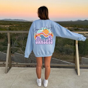 May include: A light blue sweatshirt with a graphic print of a sunset with the text "Forever Chasing Sunsets" in a rainbow font.