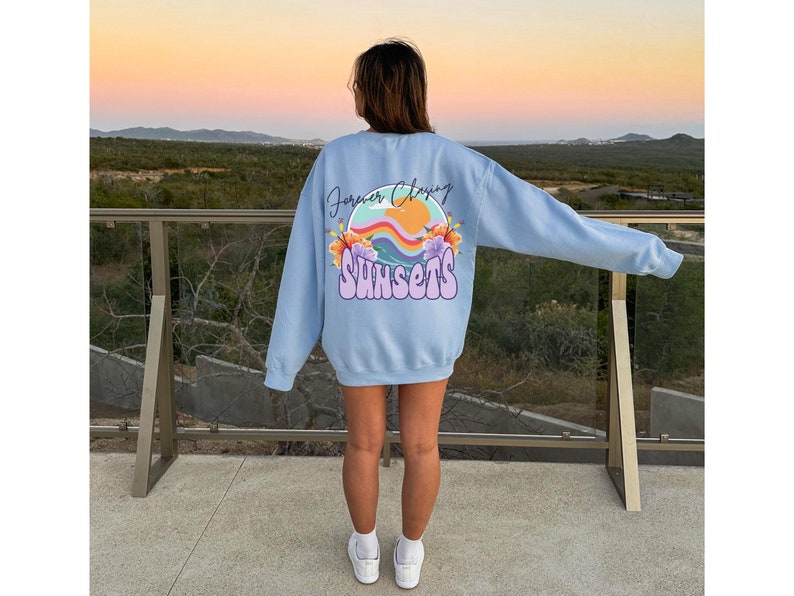 May include: A light blue sweatshirt with a graphic print of a sunset with the text "Forever Chasing Sunsets" in a rainbow font.