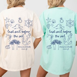 May include: Two white and light blue t-shirts with a nautical theme. The shirts feature an anchor, a ship's wheel, seashells, and the text "last sail before the veil".
