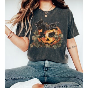 Peut inclure: Un t-shirt gris fonc&eacute; avec un motif de chats noirs &agrave; l'int&eacute;rieur d'une citrouille d'Halloween. Le motif est entour&eacute; de feuilles vertes.