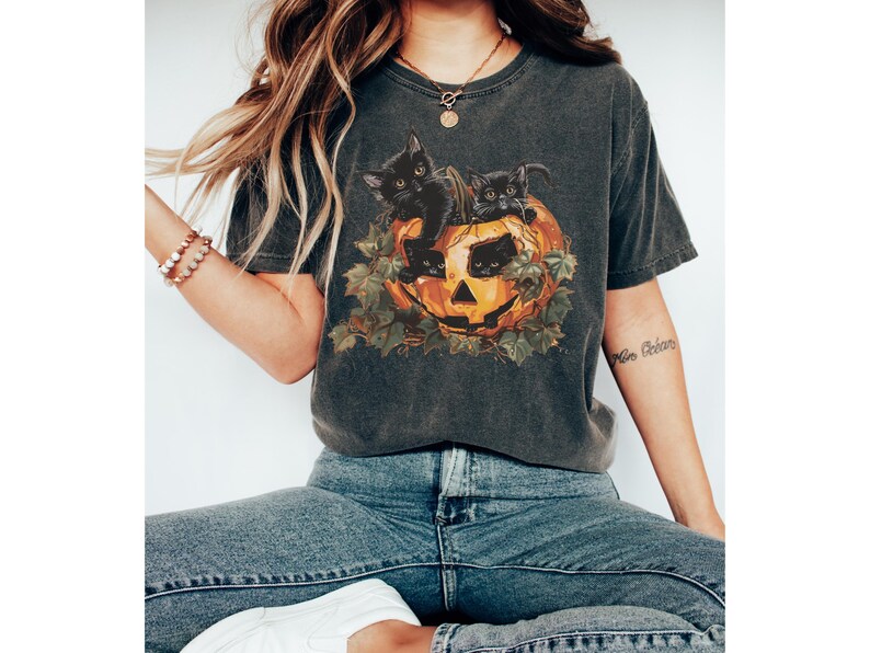 Peut inclure: Un t-shirt gris fonc&eacute; avec un motif de chats noirs &agrave; l'int&eacute;rieur d'une citrouille d'Halloween. Le motif est entour&eacute; de feuilles vertes.