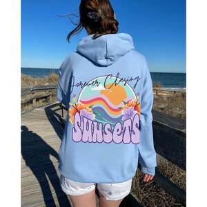 May include: A light blue hoodie featuring a graphic print of a sunset, waves, and flowers. The text "Forever Chasing Sunsets" is printed on the hoodie.