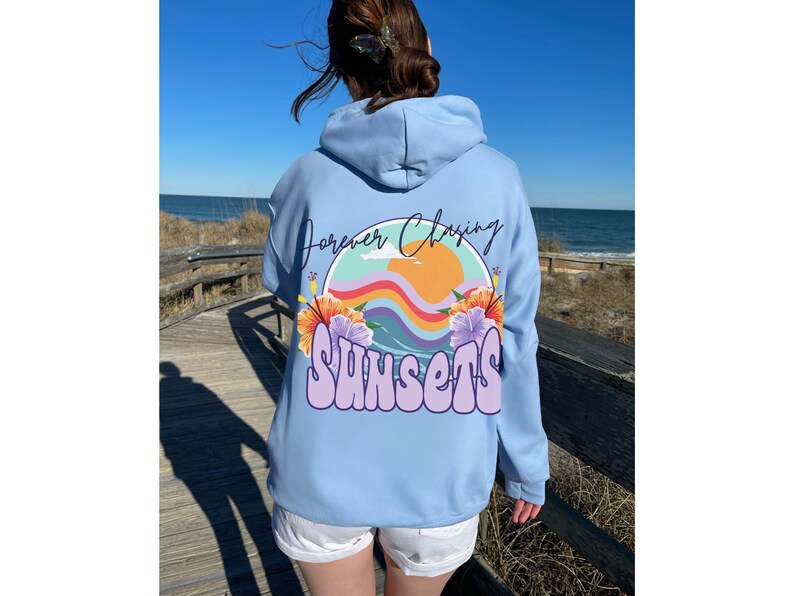 May include: A light blue hoodie featuring a graphic print of a sunset, waves, and flowers. The text "Forever Chasing Sunsets" is printed on the hoodie.
