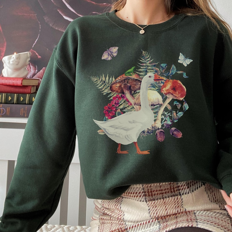 Puede incluir: Una sudadera verde oscuro con un estampado de ganso blanco y champi&ntilde;ones. El ganso est&aacute; rodeado de hojas, flores y mariposas.
