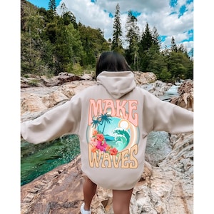 May include: Beige hoodie with a colorful graphic on the back. The graphic features a tropical scene with palm trees, waves, and flowers, along with the text "MAKE WAVES" in a retro font. The hoodie has a hood and long sleeves.