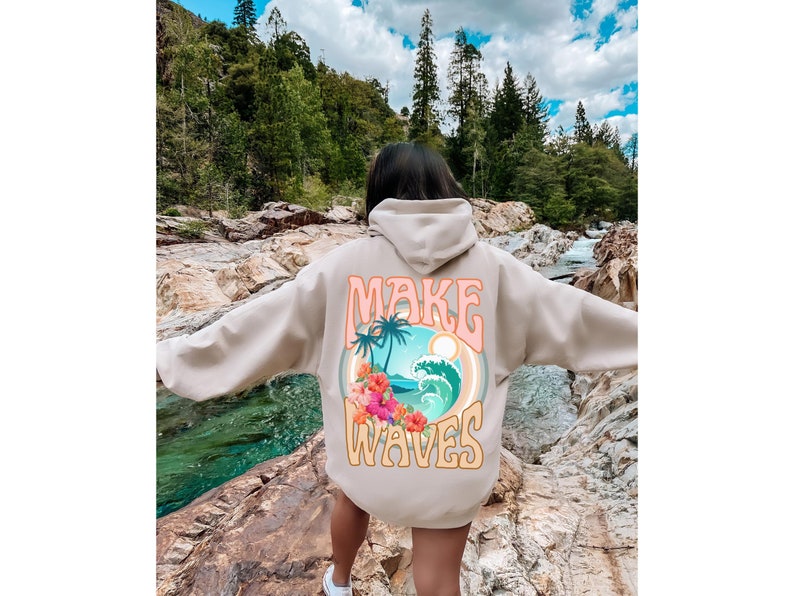May include: Beige hoodie with a colorful graphic on the back. The graphic features a tropical scene with palm trees, waves, and flowers, along with the text "MAKE WAVES" in a retro font. The hoodie has a hood and long sleeves.