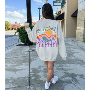 May include: A cream-coloured sweatshirt with a graphic print on the back. The graphic features a rainbow, a sun, a wave, and flowers. The text "Forever Chasing Sunsets" is written in a colourful, retro font.