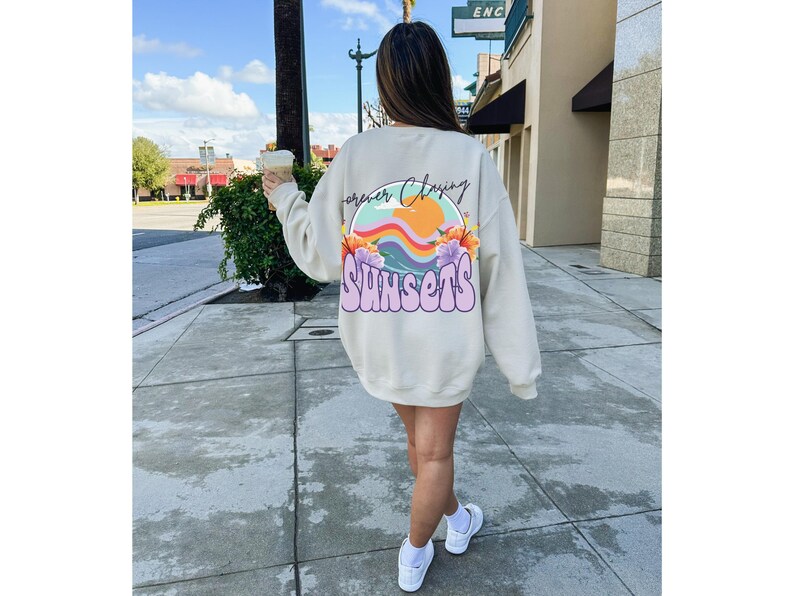 May include: A cream-coloured sweatshirt with a graphic print on the back. The graphic features a rainbow, a sun, a wave, and flowers. The text "Forever Chasing Sunsets" is written in a colourful, retro font.