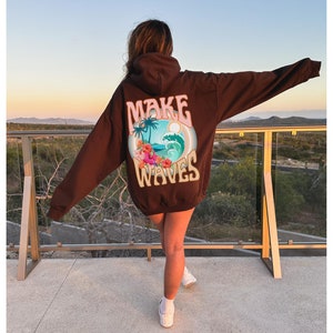 May include: A dark brown hoodie with the words "MAKE WAVES" printed on the back. The graphic includes a beach scene with palm trees, waves, and flowers. The hoodie is a relaxed fit.
