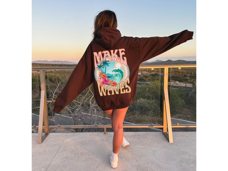 May include: A dark brown hoodie with the words "MAKE WAVES" printed on the back. The graphic includes a beach scene with palm trees, waves, and flowers. The hoodie is a relaxed fit.