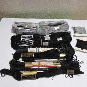 May include: A collection of embroidery floss skeins and two small cardboard spools. The skeins are primarily black and grey, featuring various paper labels including Coats & Clark's, J. & P. Coats, and Star Rayon. The threads are bundled with rubber bands.