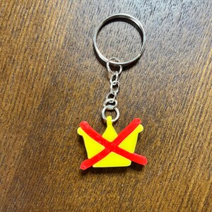 Può includere: Un portachiavi a forma di corona gialla con una "X" rossa, attaccato a una catena e un portachiavi argentati. Il portachiavi è su una superficie di legno.