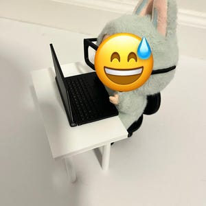 May include: A plush toy bunny wearing glasses and working on a black laptop at a small white table. The bunny is light green with pink and blue accents. The image has a white background.