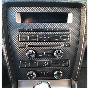 May include: A black car dashboard with a carbon fiber trim. The dashboard has a radio, climate controls, and other buttons. The radio has a display screen, buttons for tuning, and buttons for volume, seek, and sound.