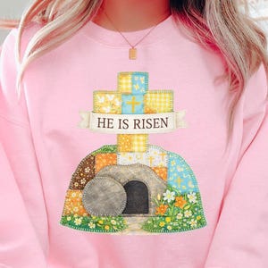 May include: A light pink sweatshirt featuring a colorful cross and tomb design. The cross is yellow and blue, with the words "HE IS RISEN" on a banner. The tomb is surrounded by flowers.