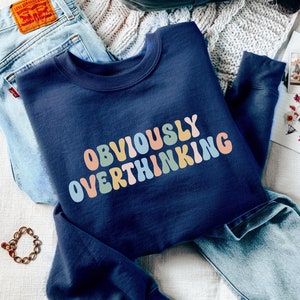 May include: Navy blue sweatshirt with the text "OBVIOUSLY OVERTHINKING" in rainbow colours. The sweatshirt is folded and laid on a white knitted blanket with a pair of blue jeans, a backpack, and a water bottle.