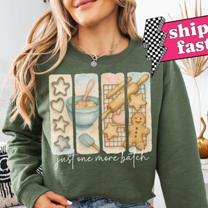 May include: A green sweatshirt with a graphic of cookie cutters, a rolling pin, a bowl of dough, and gingerbread cookies. The text "Just one more batch" is printed below the graphic.