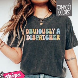 May include: A dark gray t-shirt with the text "OBVIOUSLY A DISPATCHER" in colorful retro lettering. The shirt is made of a soft, comfortable material and has a relaxed fit.