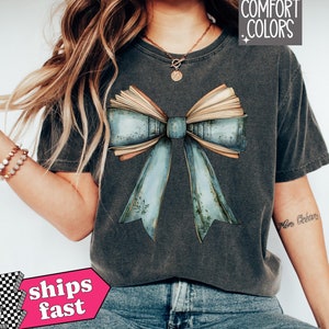 May include: Dark grey Comfort Colors t-shirt featuring a large bow design made of an open book and light blue ribbon. The bow has a vintage, distressed look. The shirt is paired with blue jeans. The words "Comfort Colors" are in the upper right corner.