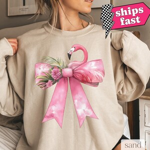 May include: A beige sweatshirt with a pink flamingo in a pink bow with floral accents. The sweatshirt has a relaxed fit and a crew neck.