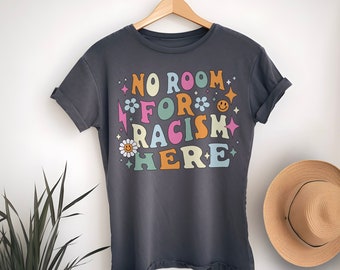 Puede incluir: Una camiseta gris oscuro con el texto "NO ROOM FOR RACISM HERE" en letras y gráficos coloridos. La camiseta tiene las mangas remangadas y está colgada de una percha de madera.
