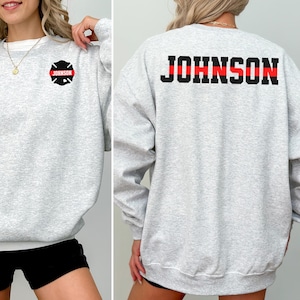 Könnte beinhalten: Hellgraues Sweatshirt mit dem Namen "JOHNSON" in schwarzen Blockbuchstaben mit einem roten Streifen in der Mitte auf dem Rücken. Auf der Vorderseite befindet sich ein schwarz-rotes Feuerwehr-Emblem mit dem Namen "JOHNSON".