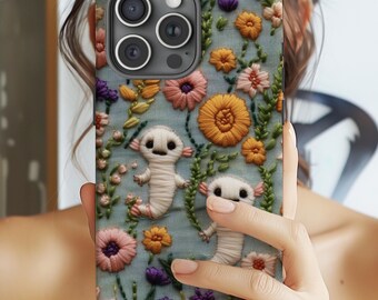 Peut inclure: Une coque de téléphone avec un motif floral mettant en scène des axolotls brodés. La coque est faite d'un matériau souple et flexible et a un dos transparent. La coque est conçue pour s'adapter à un smartphone avec un renflement de caméra.