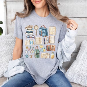 May include: A gray t-shirt featuring a watercolor illustration of books, glasses, a magnifying glass, a satchel, and other academic items. The design includes a variety of open books and stationery, creating a scholarly theme.