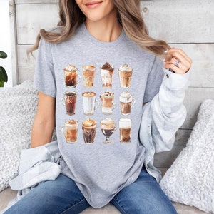 以下が含まれることがあります： 16種類の異なるコーヒー飲料のグラフィックが描かれたグレーのTシャツ。飲料はすべてガラスのカップに入っていて、ホイップクリーム、チョコレート、キャラメルなどのトッピングが施されています。