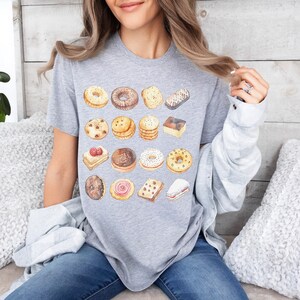 以下が含まれることがあります： 前面に様々なクッキーとペストリーがプリントされたヘザーグレーのTシャツ。クッキーとペストリーは水彩画風に描かれています。