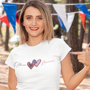 May include: White t-shirt with a graphic of two hearts. One heart is red with the Albanian flag and the other is red, white, and blue with the American flag. The text "Albanian" and "American" are written on either side of the hearts.