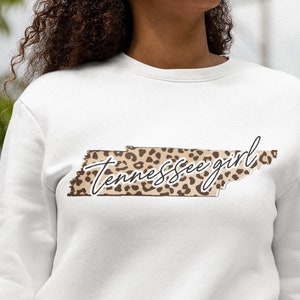 Puede incluir: Sudadera blanca con un gráfico del estado de Tennessee en un diseño de estampado de leopardo. El texto "Tennessee girl" está escrito en cursiva sobre el contorno del estado.