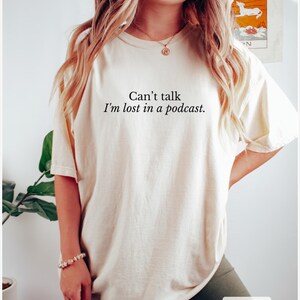 May include: A cream colored t-shirt with the text "Can't talk I'm lost in a podcast." printed in black.