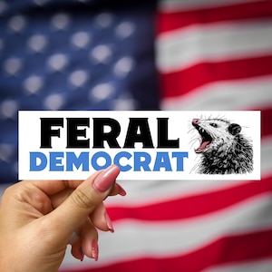Peut inclure: Un autocollant blanc avec le texte "FERAL DEMOCRAT" en lettres noires et une illustration en noir et blanc d'un opossum la gueule ouverte. L'autocollant est tenu par une main sur un fond flou d'un drapeau américain.