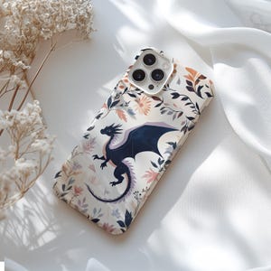May include: A white phone case with a floral design and a black dragon illustration. The phone case has a camera lens cutout and a white background.