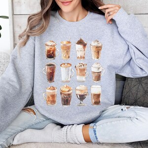 May include: A grey sweatshirt featuring a watercolour design of 12 different coffee drinks. The coffee drinks are arranged in a grid pattern and include lattes, cappuccinos, macchiatos, and other coffee beverages.