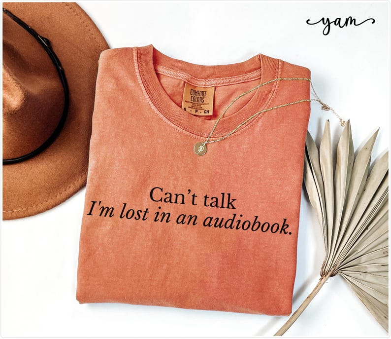 May include: A coral-colored t-shirt with the text "Can't talk I'm lost in an audiobook." printed in black.