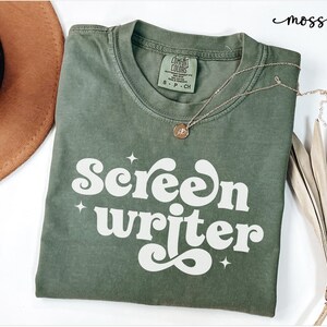 May include: Olive green t-shirt with the words "Screen Writer" in white, stylized font. A gold necklace with a pendant rests on the shirt. A brown hat is visible in the upper left corner.