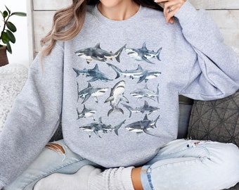 May include: A pale grey sweatshirt displaying a watercolour illustration of diverse shark species. The sharks are rendered in shades of grey, blue, and white, creating a detailed and artistic design. The sweatshirt is a comfortable, casual style.