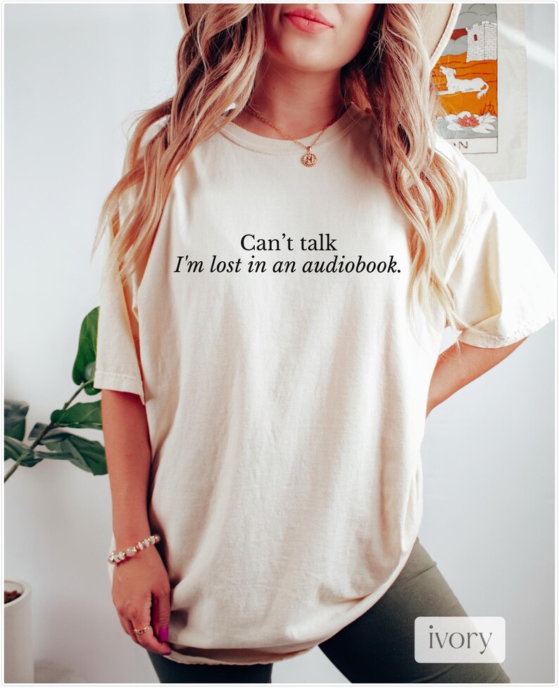 May include: A woman wearing a light beige t-shirt with the text "Can't talk. I'm lost in an audiobook." printed on it. The shirt is a relaxed fit and has a crew neck.