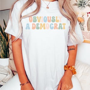 Puede incluir: Camiseta blanca con el texto "OBVIOUSLY A DEMOCRAT" en letras redondas y de colores.