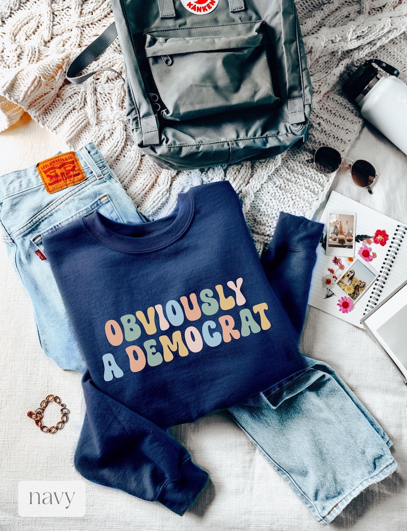 May include: A navy blue sweatshirt with the text "OBVIOUSLY A DEMOCRAT" in rainbow-colored letters. The sweatshirt is folded and laid on a white knitted surface with blue jeans and a gray backpack.